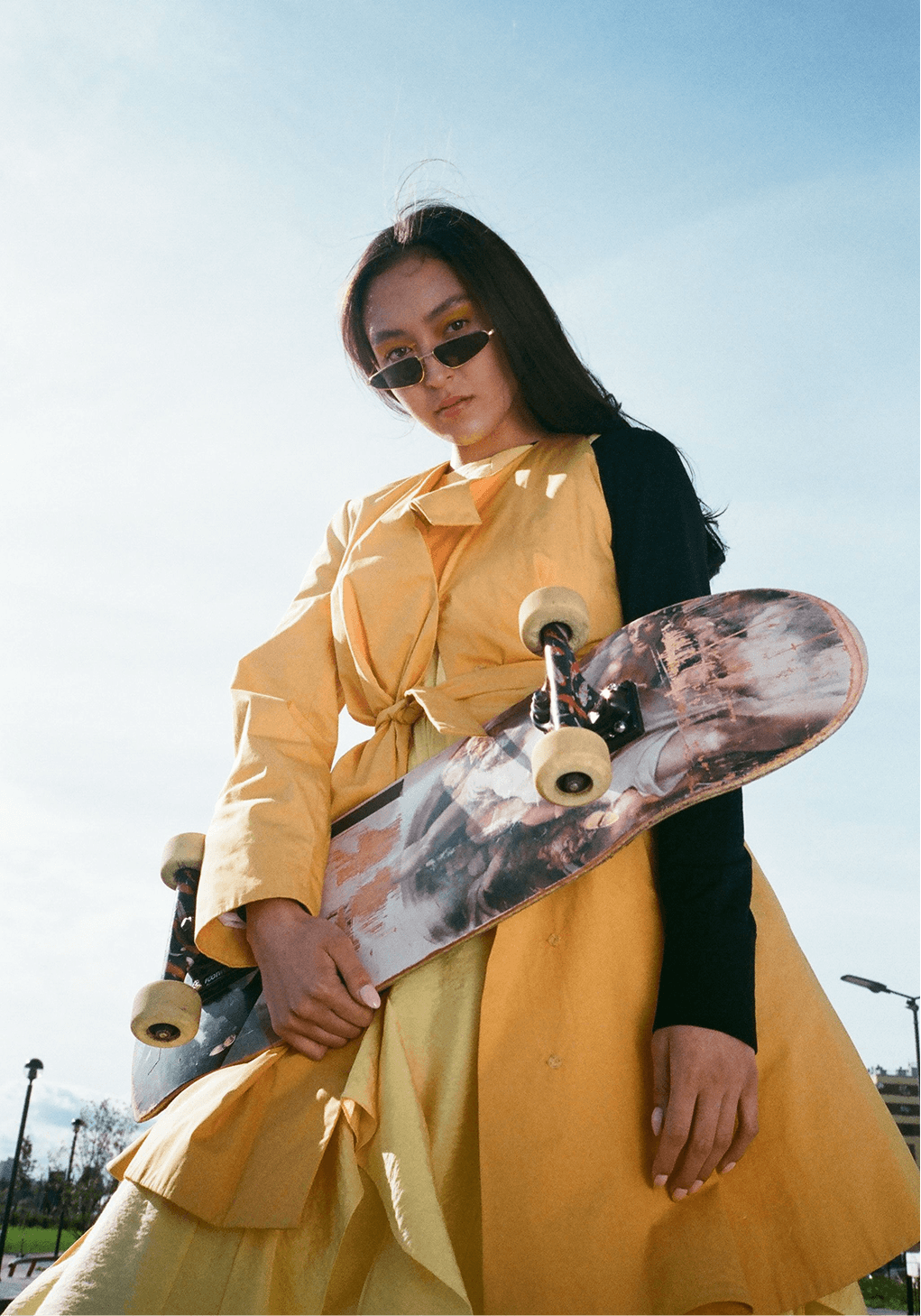 A young person in a yellow coat and sunglasses holding a skateboard against an open sky