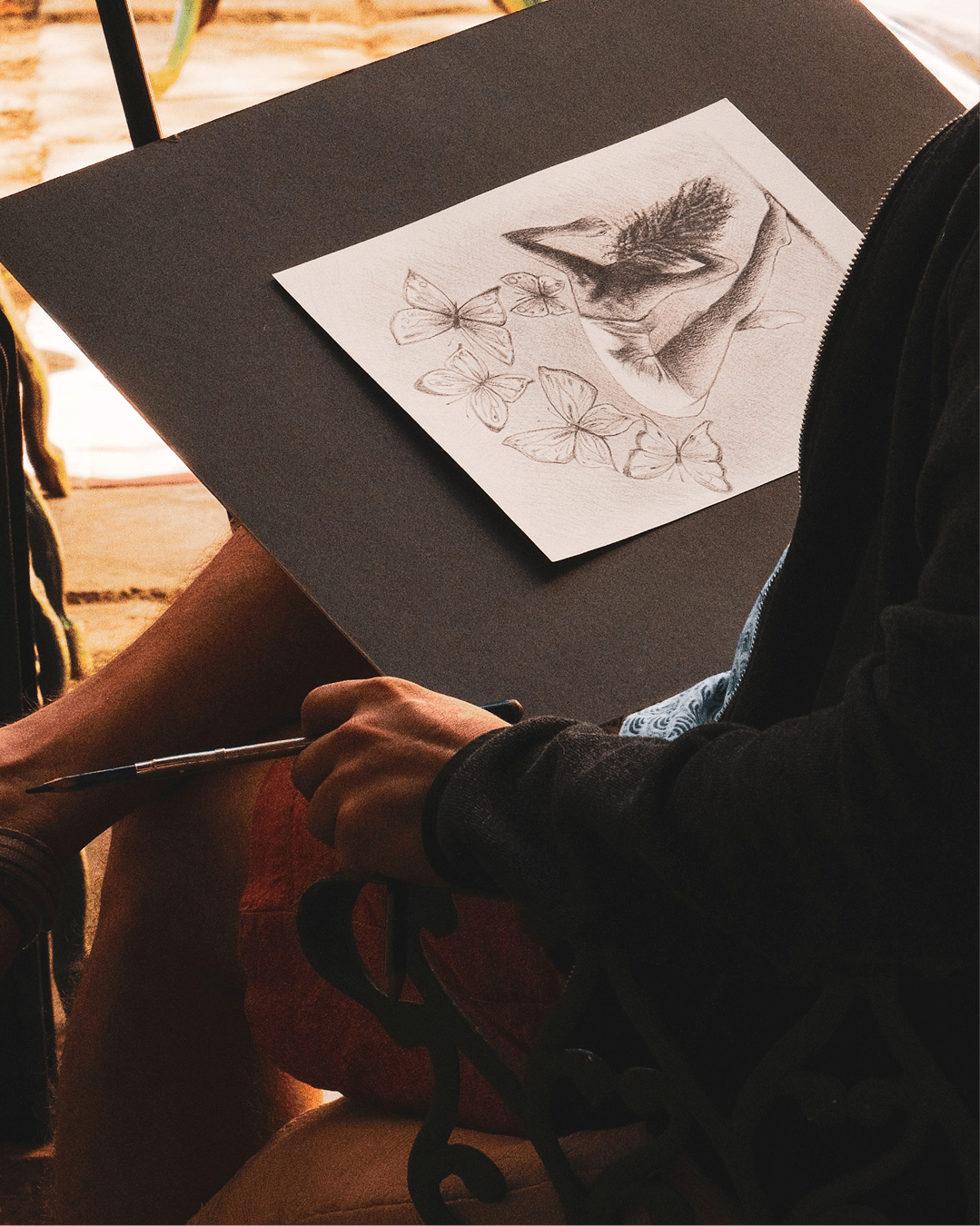 A person sketching a whale and butterflies in pencil, seated outdoors in warm evening light